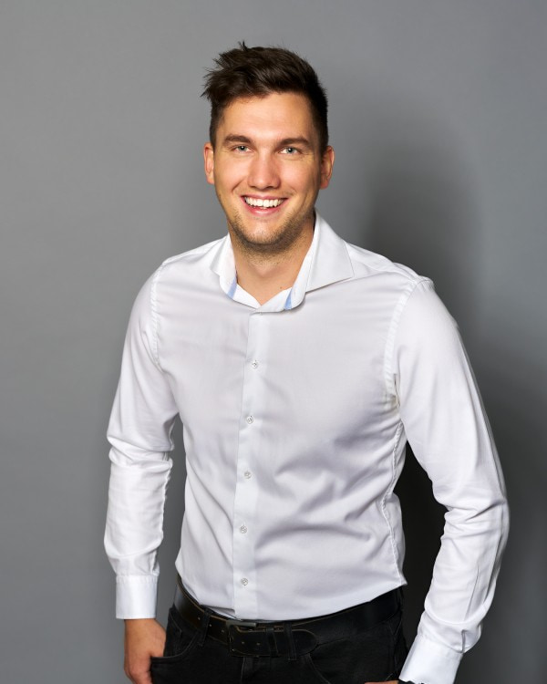 Patrick Haidacher; -	Der Mitarbeiter für die Technische Leitung am Standort in Österreich trägt ein weißes Hemd, hat seine Haltung leicht nach vorne gebeugt und lacht mit breitem Mund in die Kamera.