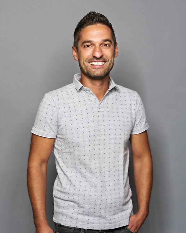 Rene Greiderer; -	Ein Mitarbeiter für Logistik und Disposition hat ein hellgraues Poloshirt mit Muster an, trägt seine kurzen Haare nach oben und hat einen Arm in der Hosentasche eingesteckt.