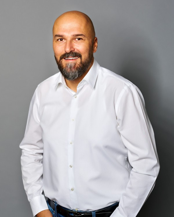 Gerold Huber; -	Einer von zwei Geschäftsführern, der ein weißes Hemd trägt und vor einem grauen Hintergrund steht, trägt einen Vollbart und hat eine Glatze.